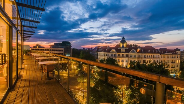 Un balcón con vista a un paisaje urbano al atardecer, que muestra edificios y una estructura en forma de cúpula.