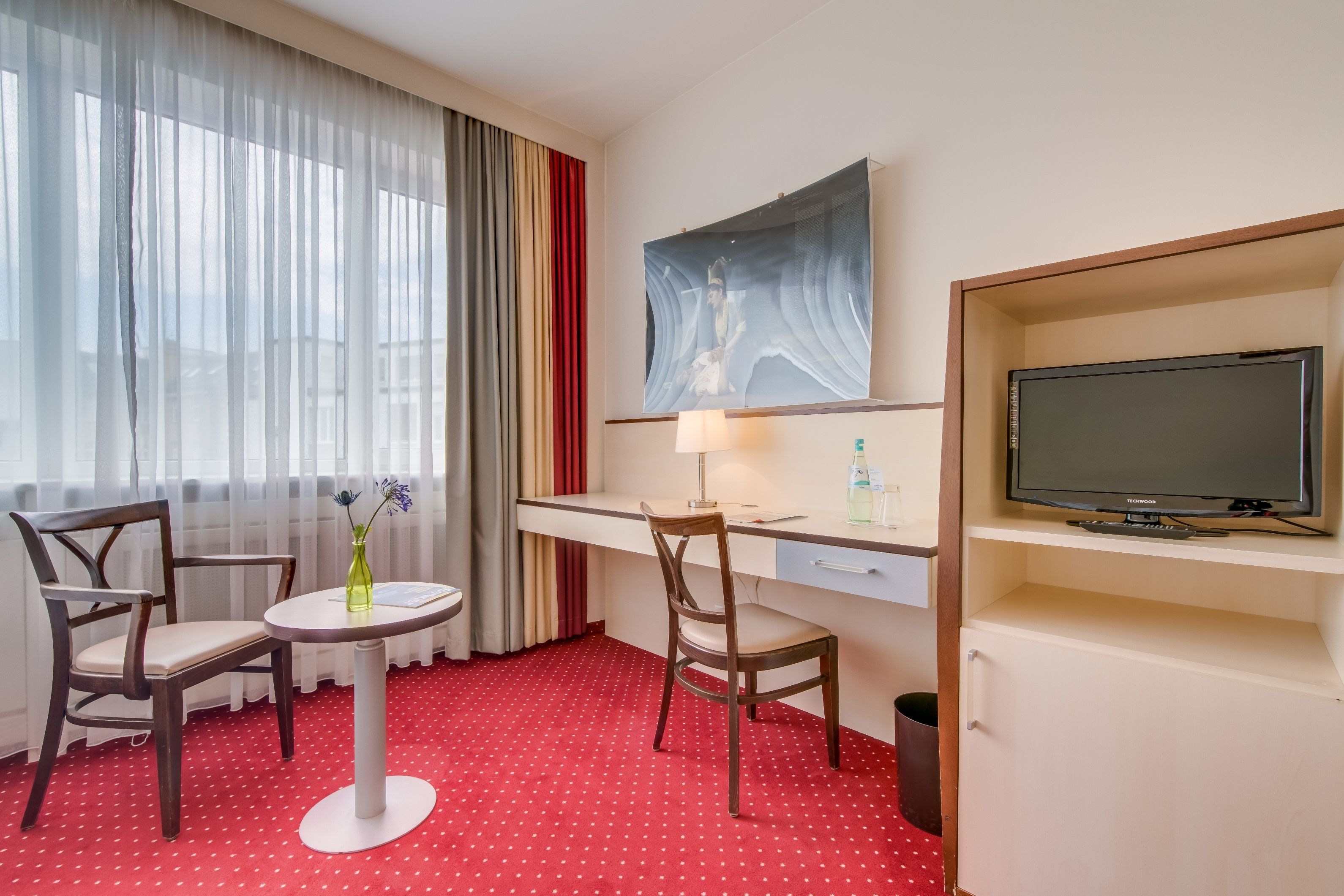 A hotel room with a desk, two chairs, a TV, a painting, and a window with curtains.