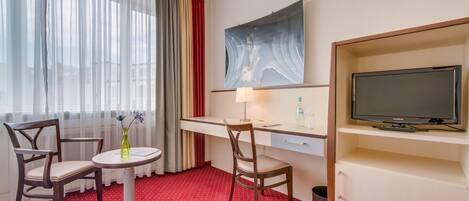 A hotel room with a desk, two chairs, a TV, a painting, and a window with curtains.