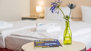 Una habitación de hotel con una cama, una mesita de noche, una lámpara, una revista y un jarrón con flores.