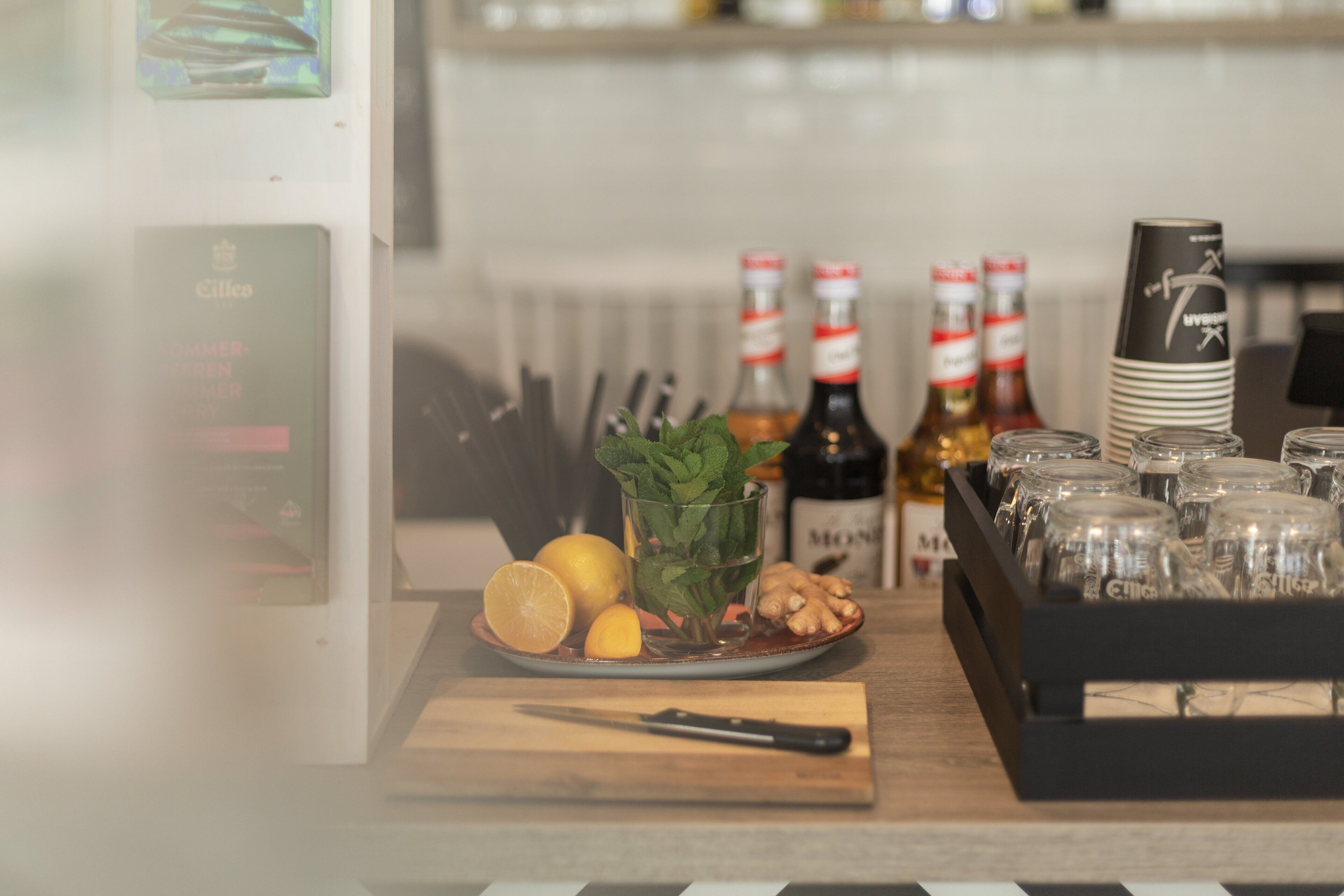 A kitchen counter with a cutting board, a knife, a plate with lemons, mint leaves, and ginger. There are bottles of alcohol and stacked glasses.