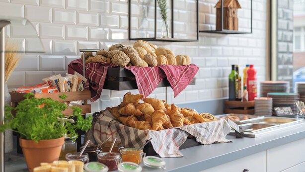A breakfast buffet with pastries and bread, a potted plant, and a window with a brick wall. - Garner Hotel Berlin - Wilmersdorf by IHG (Berlin)
