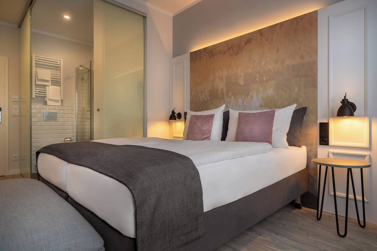 A modern hotel room with a large bed, bedside tables, a glass shower enclosure, and a view of the outside.