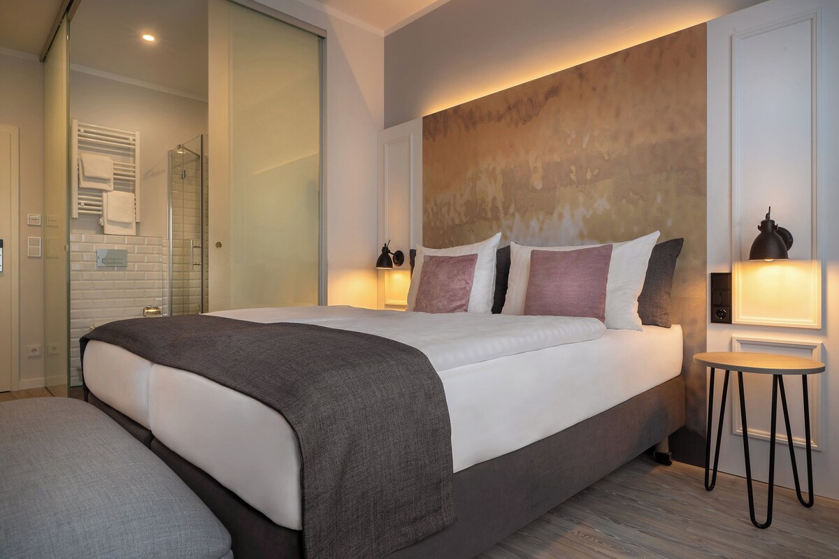 Habitación de hotel moderna con una cama grande, mesitas de noche, cabina de ducha de vidrio y vistas al exterior.