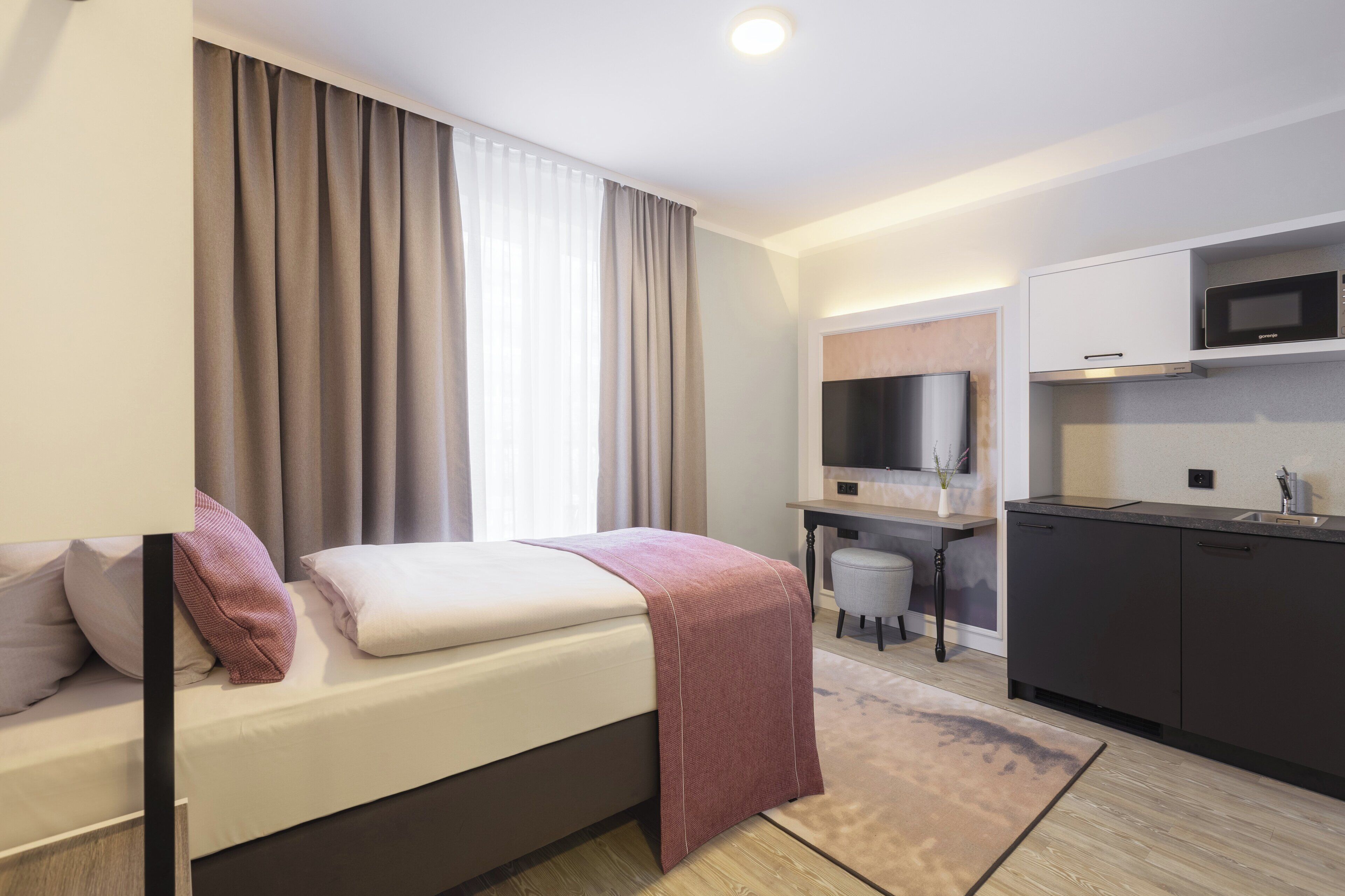 A modern hotel room with a bed, a desk, a television, and a kitchenette.