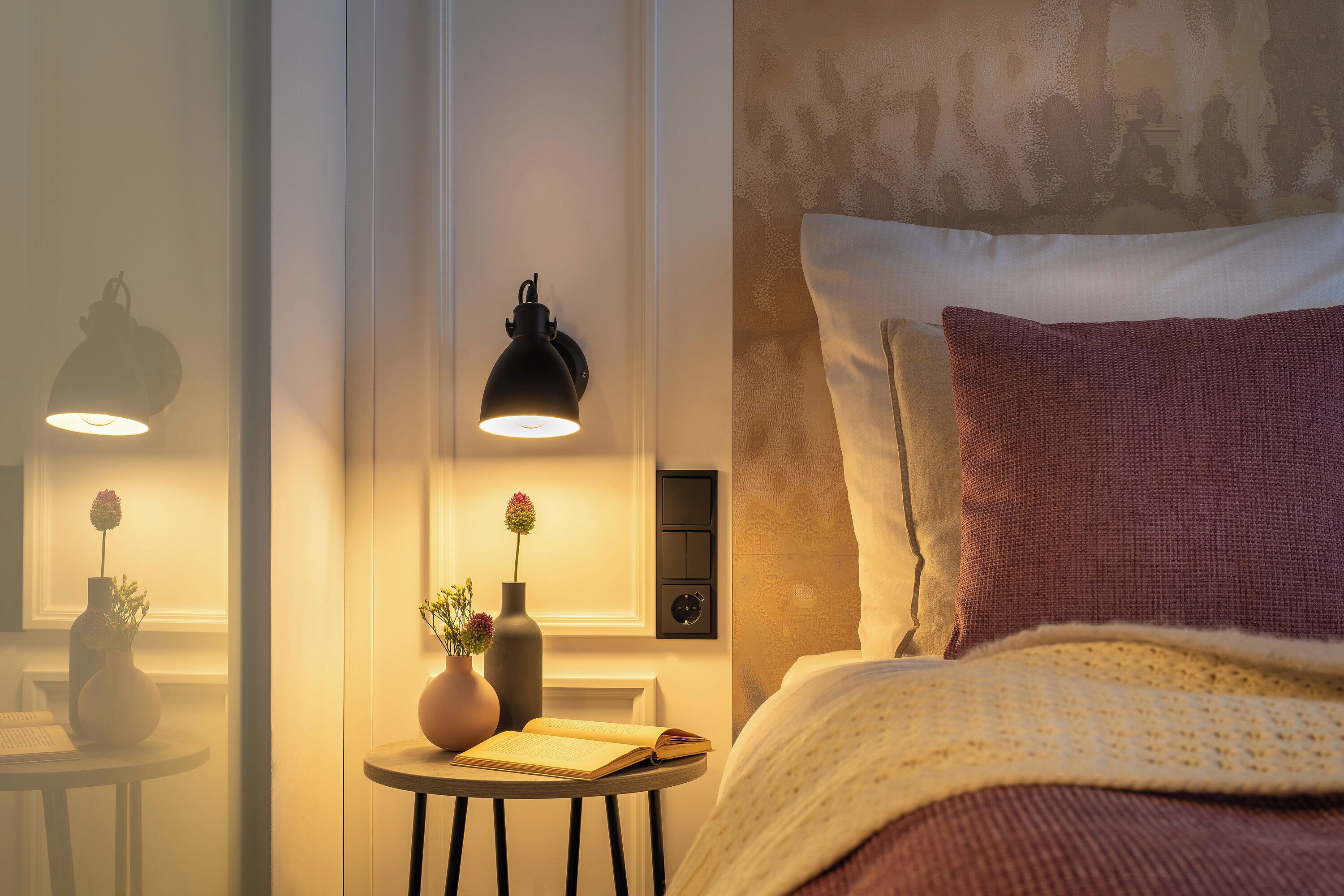 A bedroom with a bed, a bedside table with a book and a vase, a wall-mounted lamp, and a thermostat.