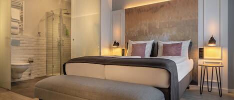 A modern bedroom with a large bed, bedside tables, a bench, a glass-enclosed shower, and a bathroom with a toilet and sink.