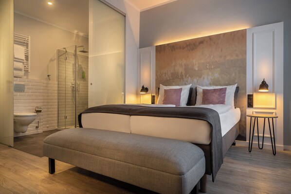 A modern bedroom with a large bed, bedside tables, a bench, a glass-enclosed shower, and a bathroom with a toilet and sink. - Garner Hotel Berlin - Wilmersdorf by IHG (Berlin)