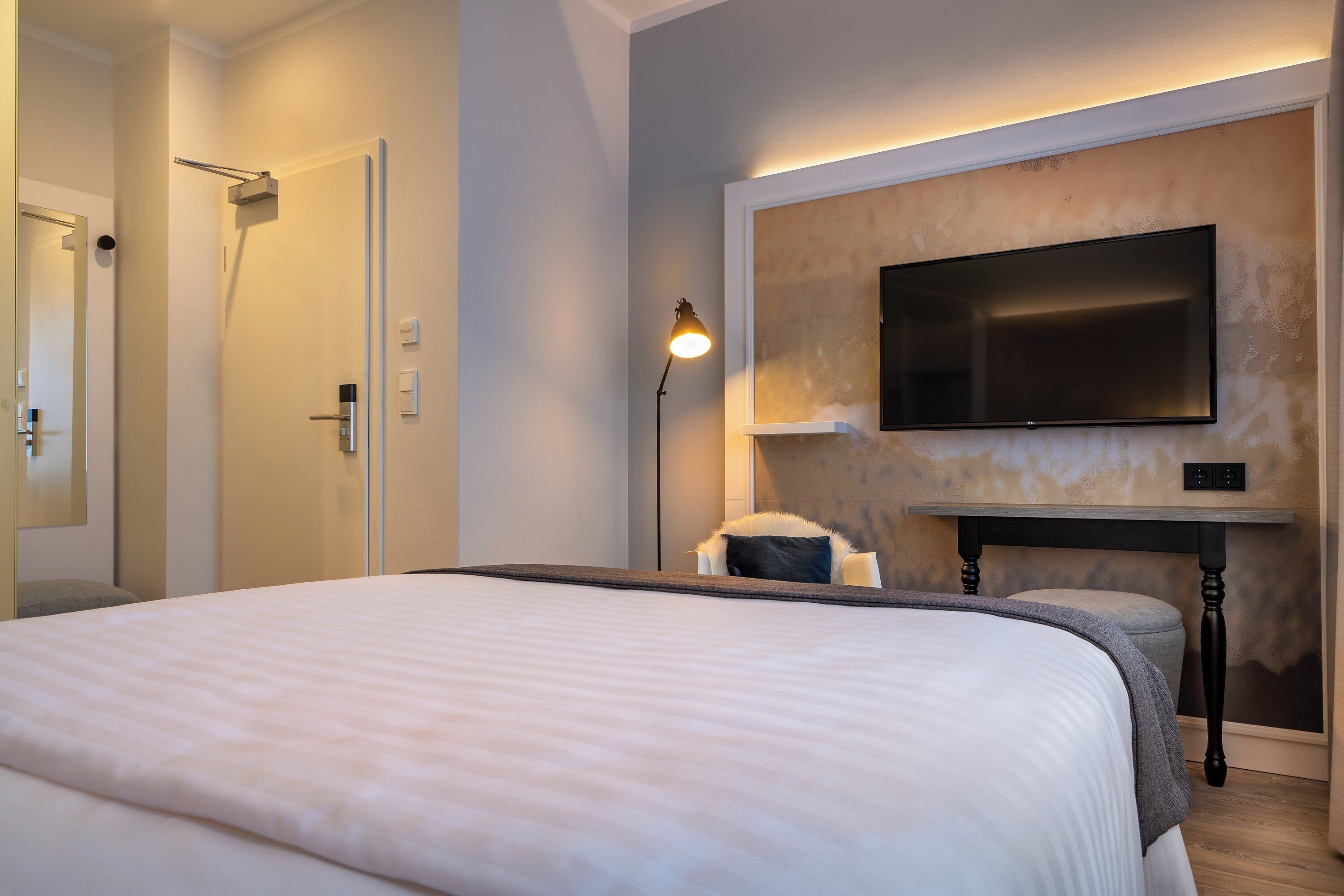 A modern hotel room with a large bed, a flat-screen TV mounted on the wall, a bedside table, and a nightstand.