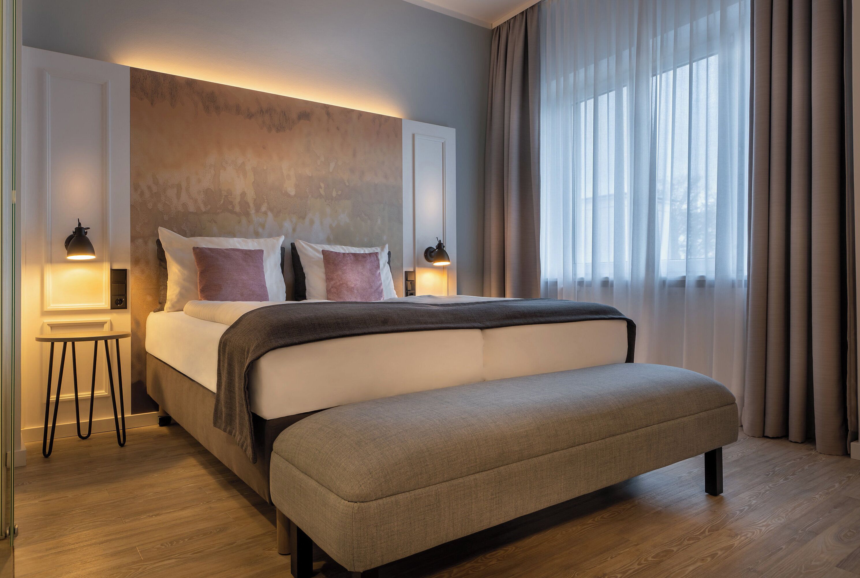 Une chambre à coucher moderne, dotée d’un grand lit, de lampes de chevet et offrant une vue sur l’extérieur à travers de fines rideaux.