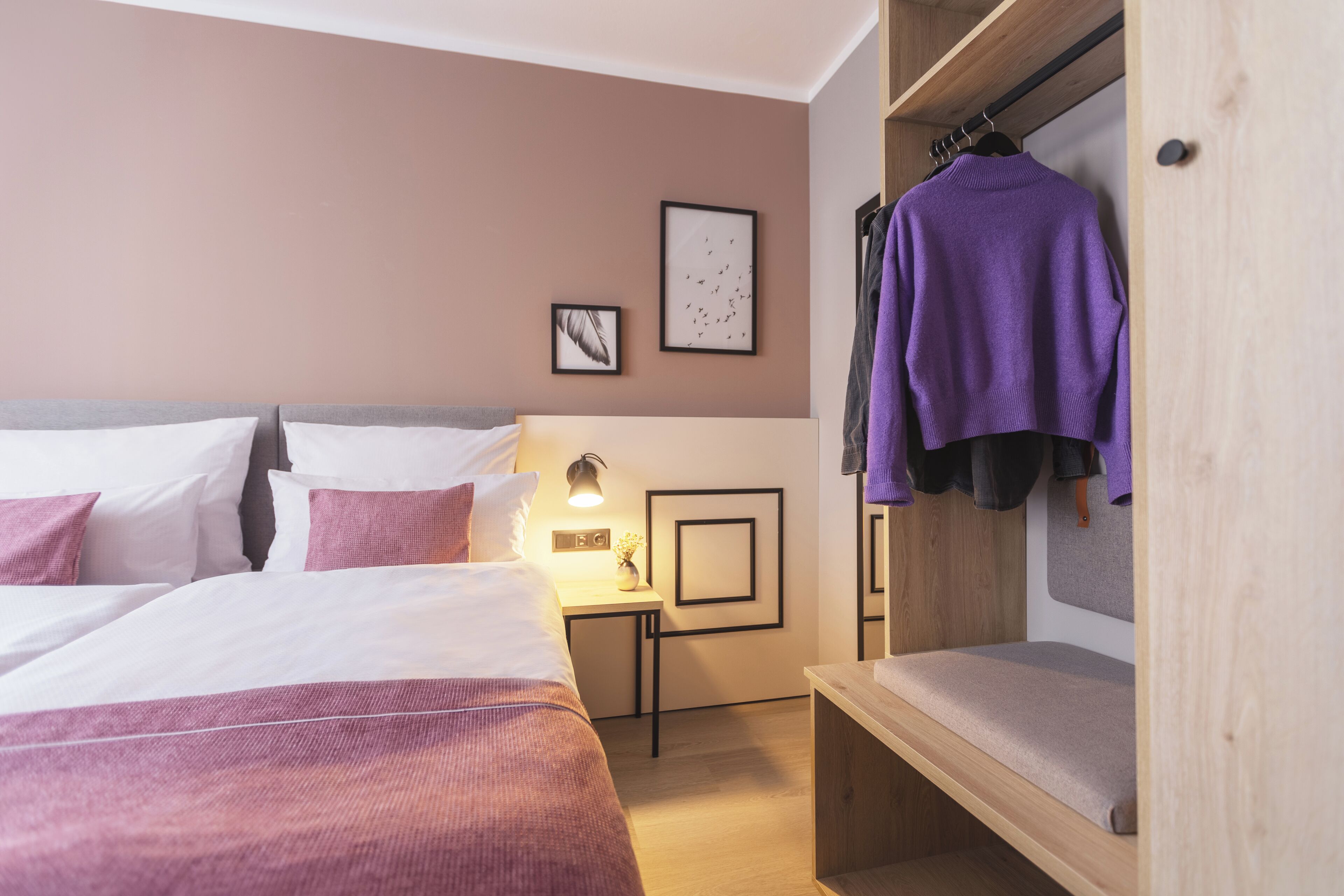 A hotel room with a large bed, a bedside table with a lamp, and a wardrobe with hanging clothes.