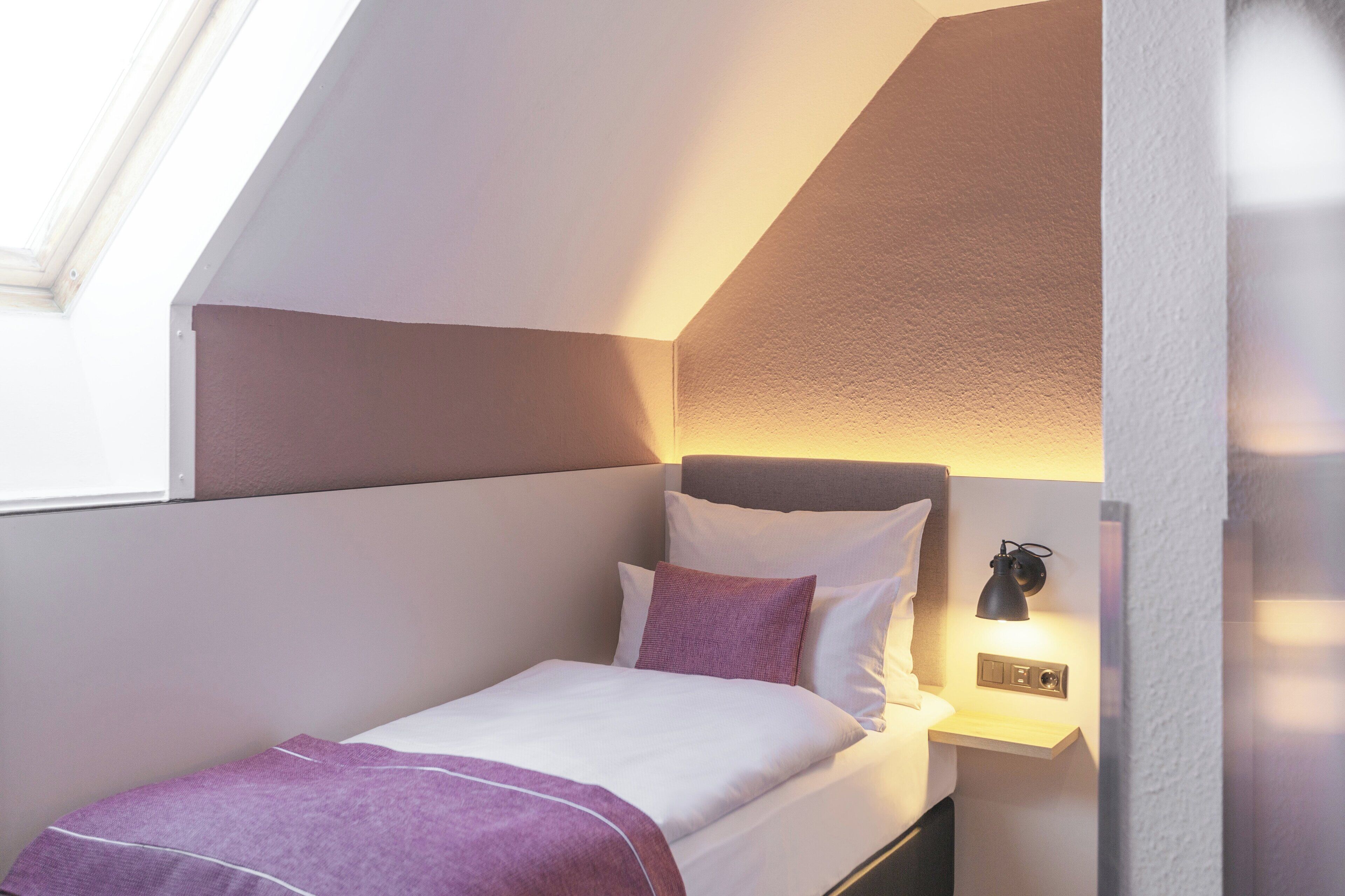 A small, neatly made bed with a headboard, a bedside table with a lamp, and a window providing natural light.