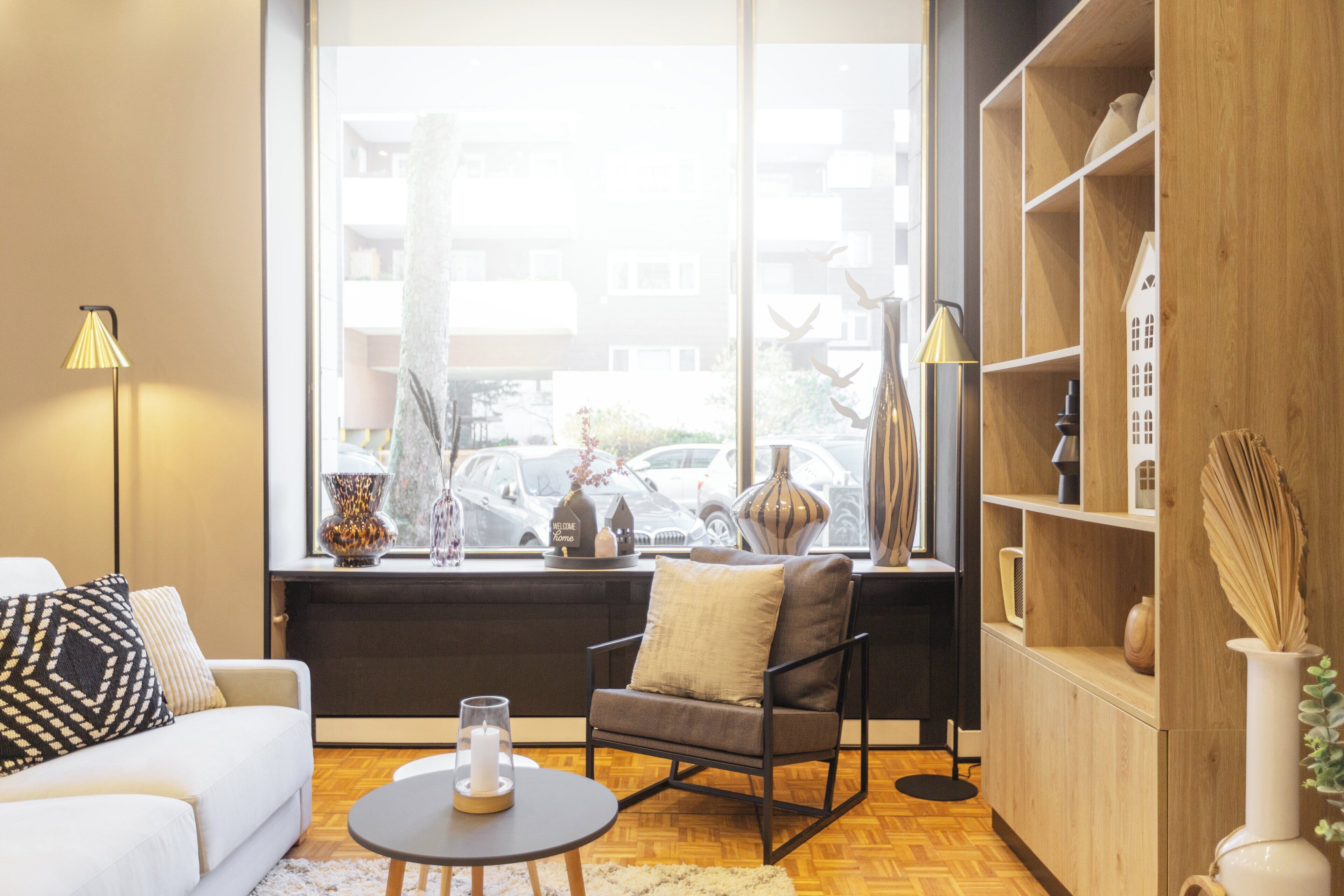 En moderne stue med sofa, lænestol og sofabord, store vinduer med udsigt til parkerede biler, og en reol i træ.