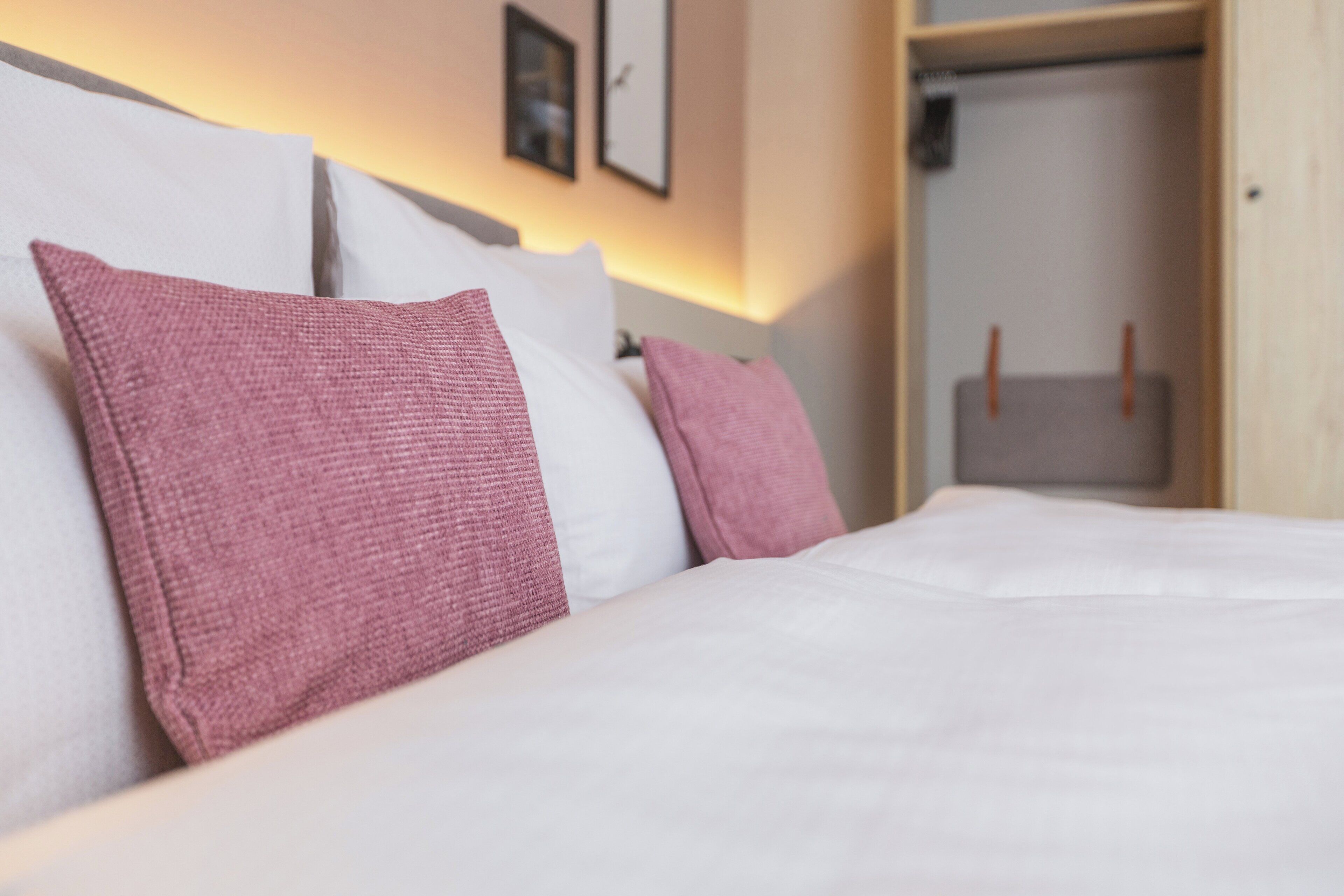 Una cama bien hecha con almohadas blancas y rosas, un cabecero de madera y un armario con un bolso colgado.