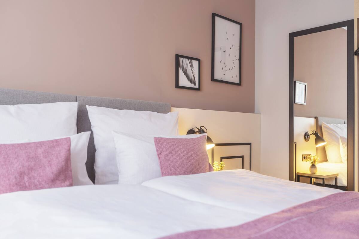 A neatly made bed with white and pink bedding, a bedside table with a lamp, and framed artwork on the wall.