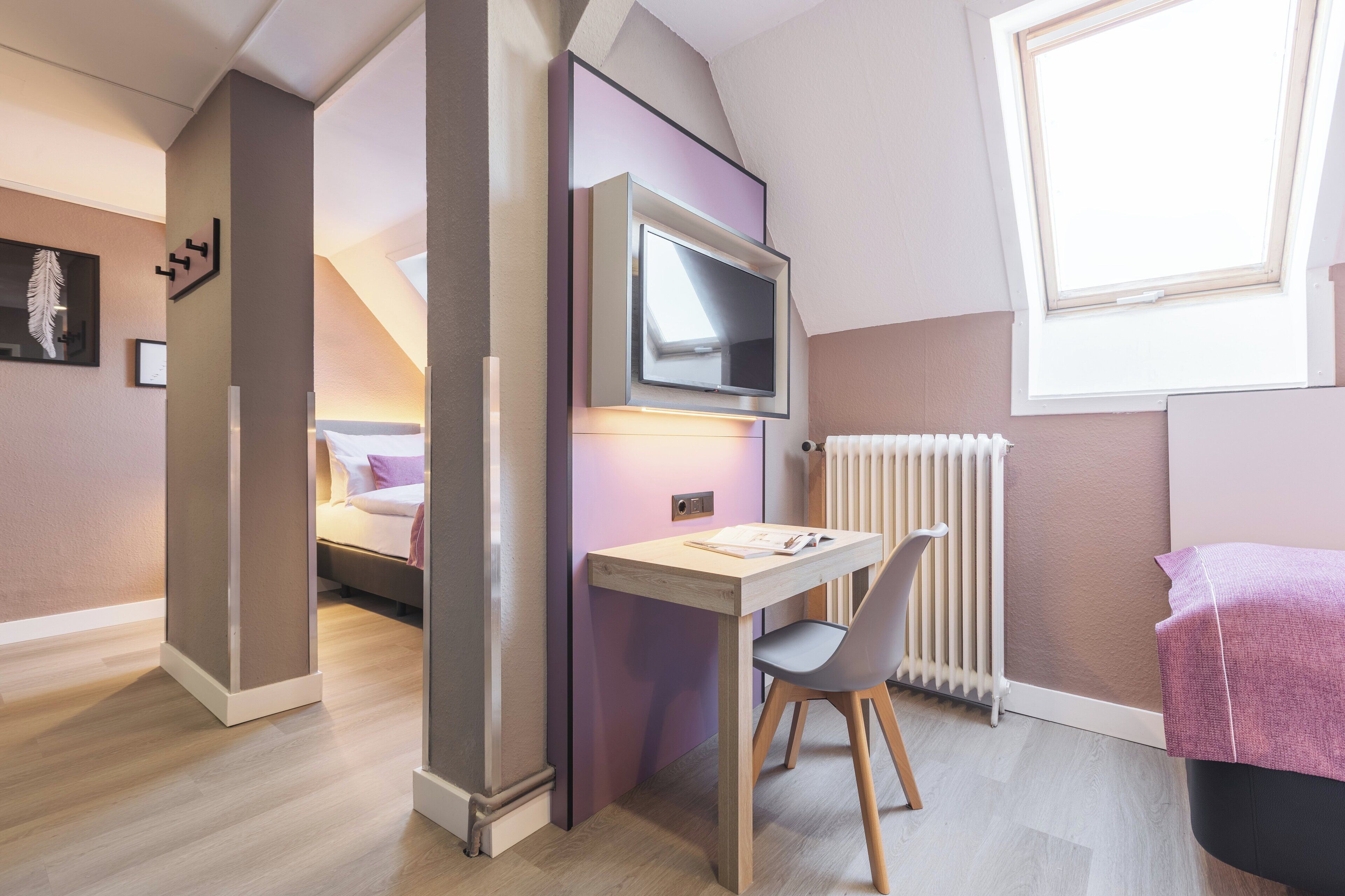 A modern bedroom with a bed, a desk with a chair, a TV mounted on the wall, and a window providing natural light.
