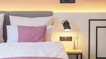 Ein ordentlich bezogenes Bett mit weißen und rosa Kissen, einer Nachttischlampe und zwei Vasen mit Blumen auf einem kleinen Tisch daneben.