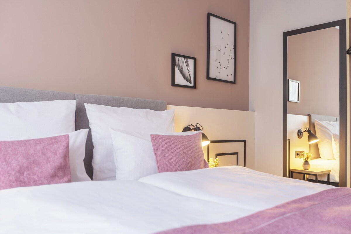 Una cama bien hecha con ropa de cama blanca y rosa, una mesita de noche con una lámpara y cuadros en la pared.