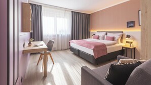 A modern hotel room with a bed, bedside table, lamp, and a view of the city through large windows.