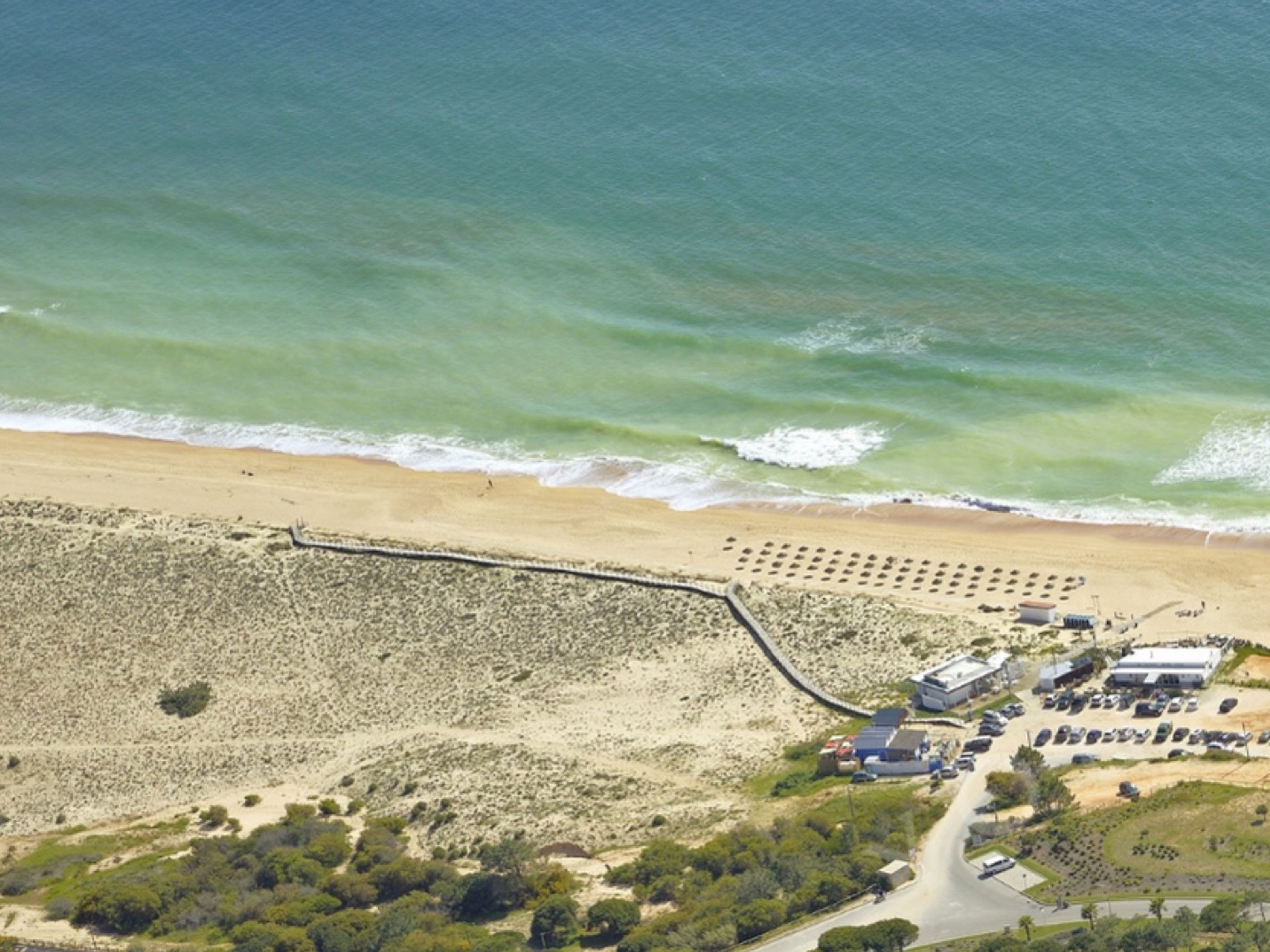 Eine Luftaufnahme eines Strandes mit einer langen Mole, die ins Meer führt, einem Parkplatz und Gebäuden in der Nähe.