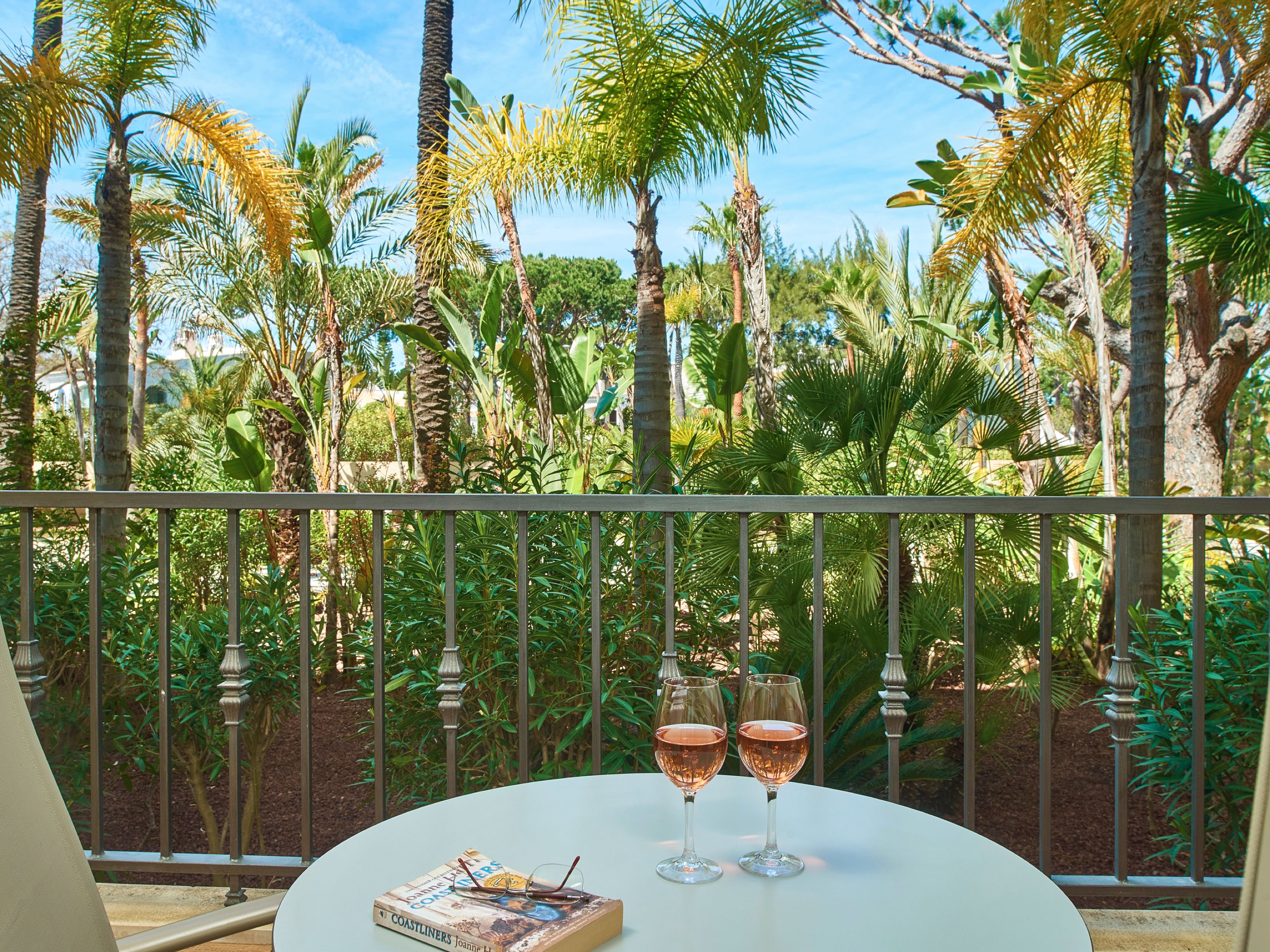 A balcony with two wine glasses and a book on a table, surrounded by lush greenery and palm trees.