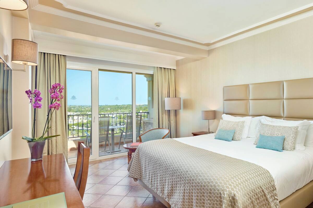 A hotel room with a large bed, a desk, a chair, a balcony with a view, and a potted plant.