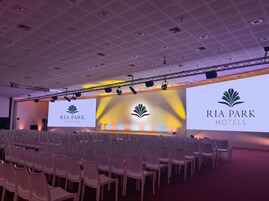 Un salón de conferencias con hileras de sillas blancas, un escenario con una pantalla grande mostrando el logotipo de RIA PARK HOTELS, e iluminación empotrada y una estructura de soporte en el techo.