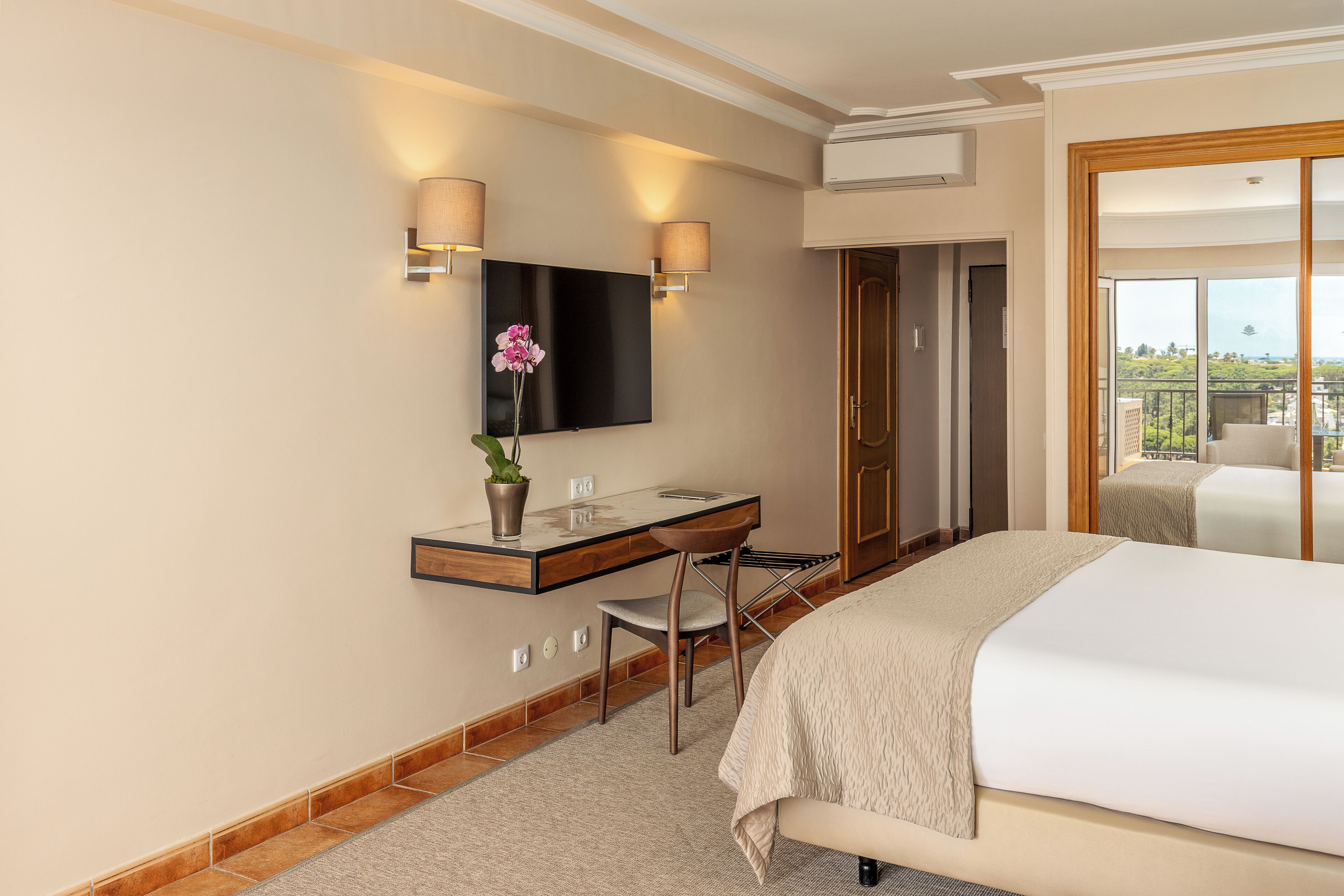 A hotel room with a bed, a desk with a chair, a flat-screen TV mounted on the wall, and a view of the outdoors.