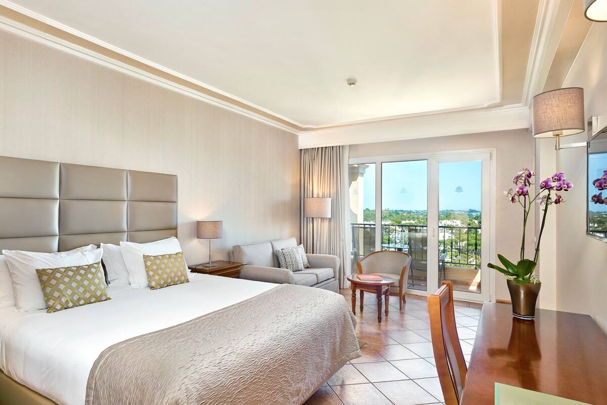 A hotel room with a large bed, a sofa, a chair, a small table, and a balcony with a view.