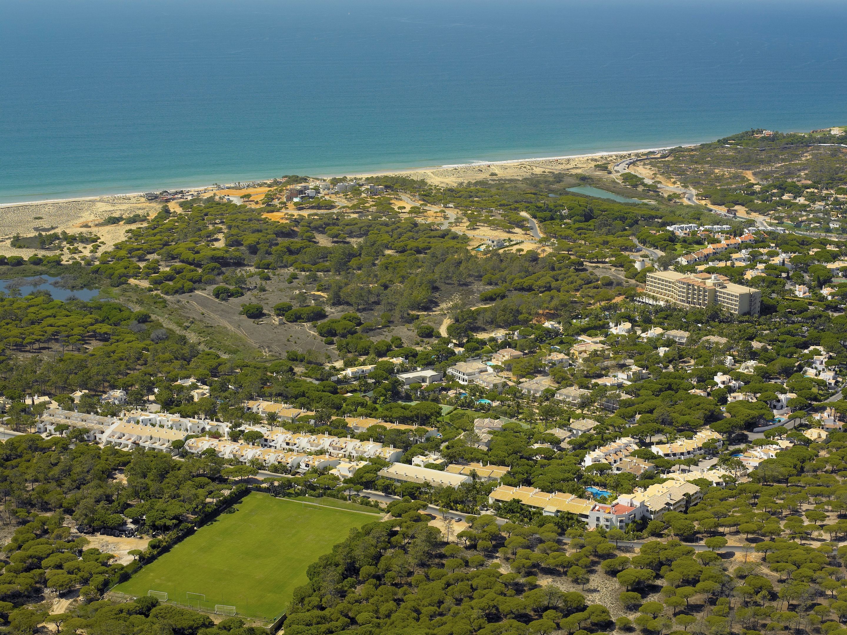 An aerial view of a coastal residential area with a beach, dense greenery, and a large building complex.