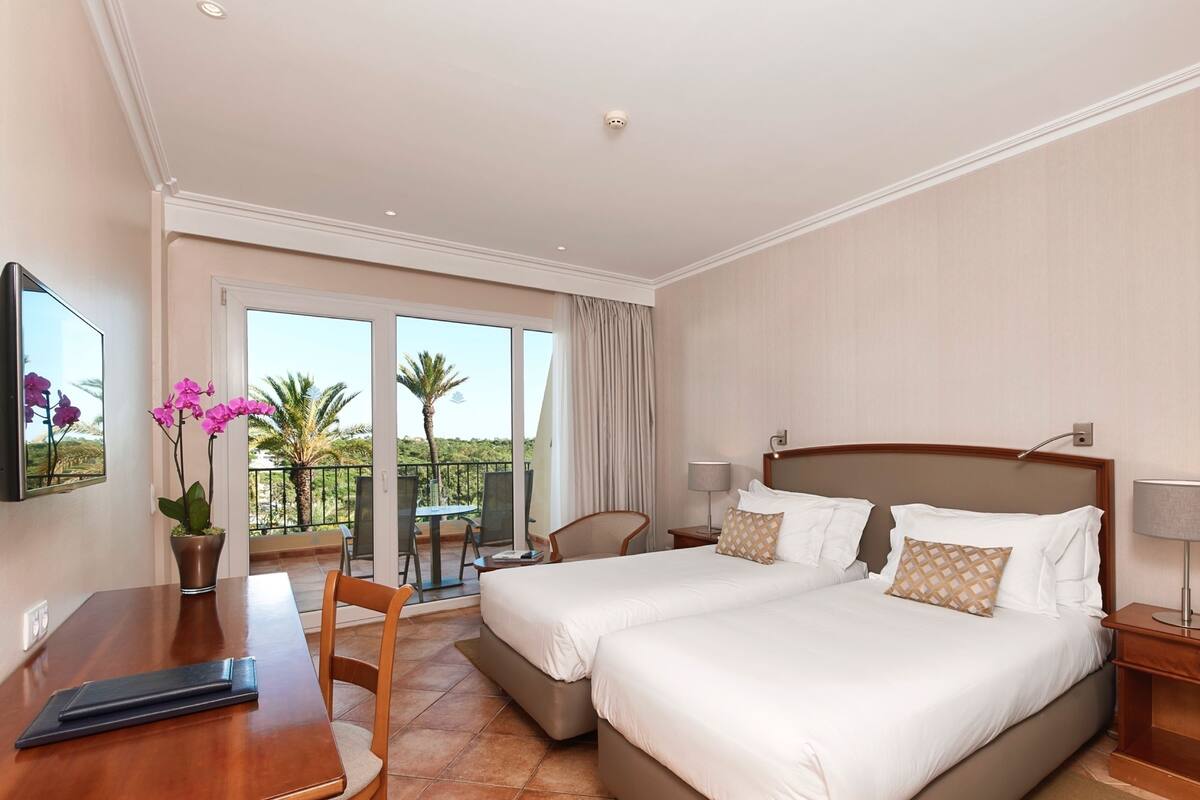 A hotel room with a bed, two bedside tables, a desk, a chair, a potted plant, and a balcony with a view of palm trees.