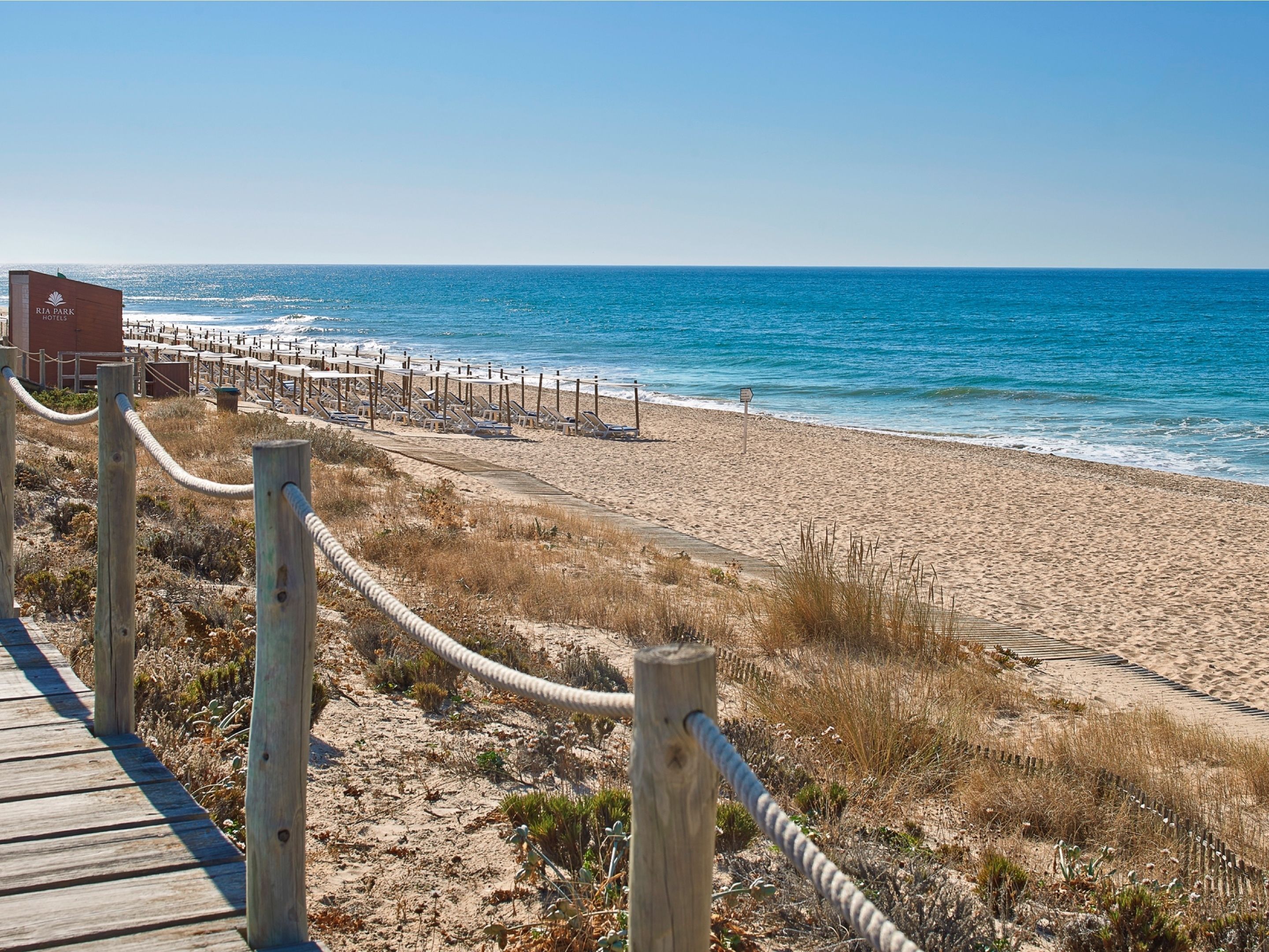 A beach with a wooden boardwalk leading to a row of sun loungers and a sign for RIA PARK HOTELS.
