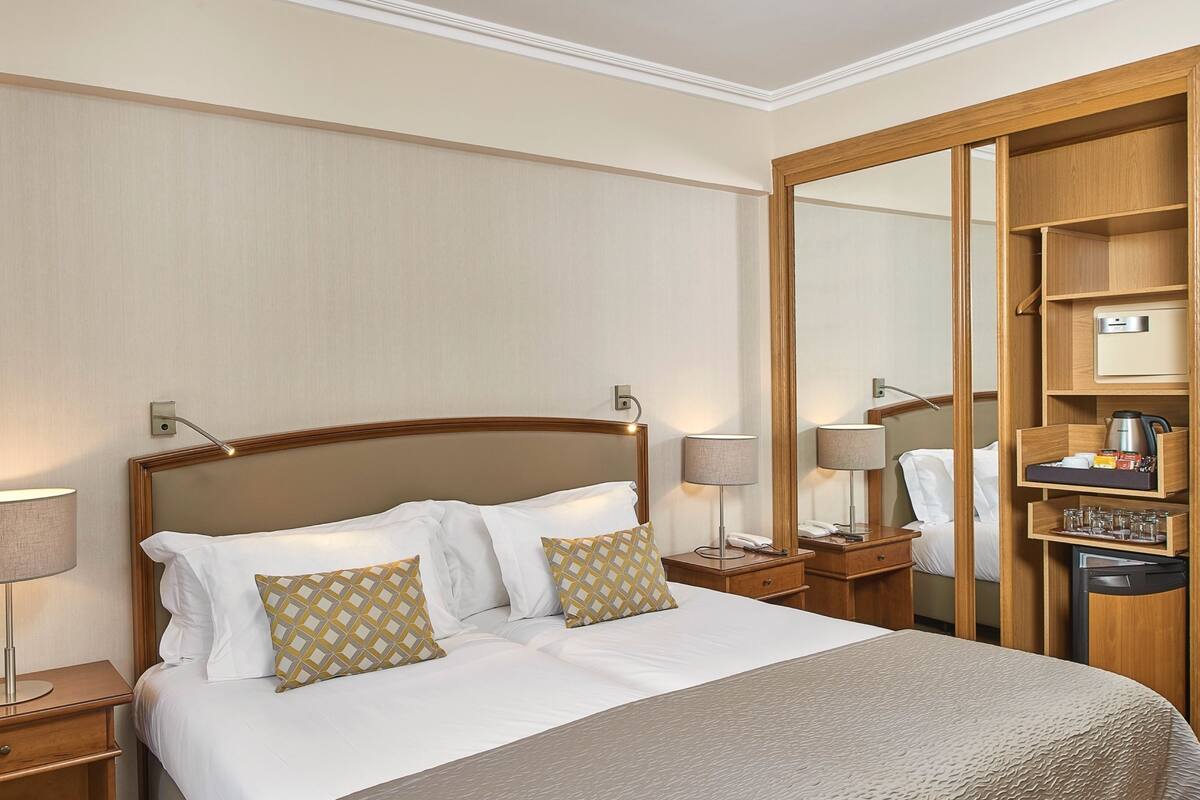 A hotel room with a large bed, wooden headboard, bedside tables with lamps, a mirror, and a wardrobe with shelves.