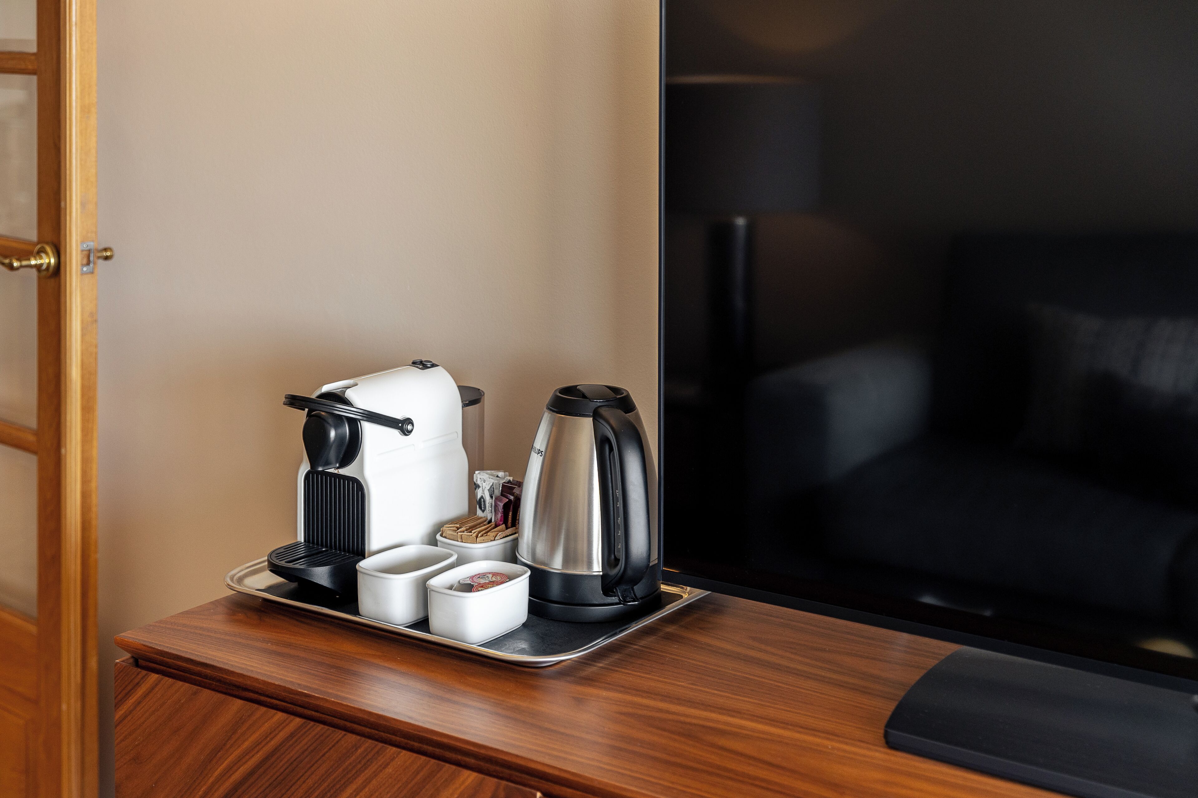 A hotel room with a wooden nightstand holding a coffee maker, a kettle, and a tray with cups and a box of tissues.