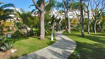 A paved pathway curving through a well-maintained garden with various trees and a building in the background.