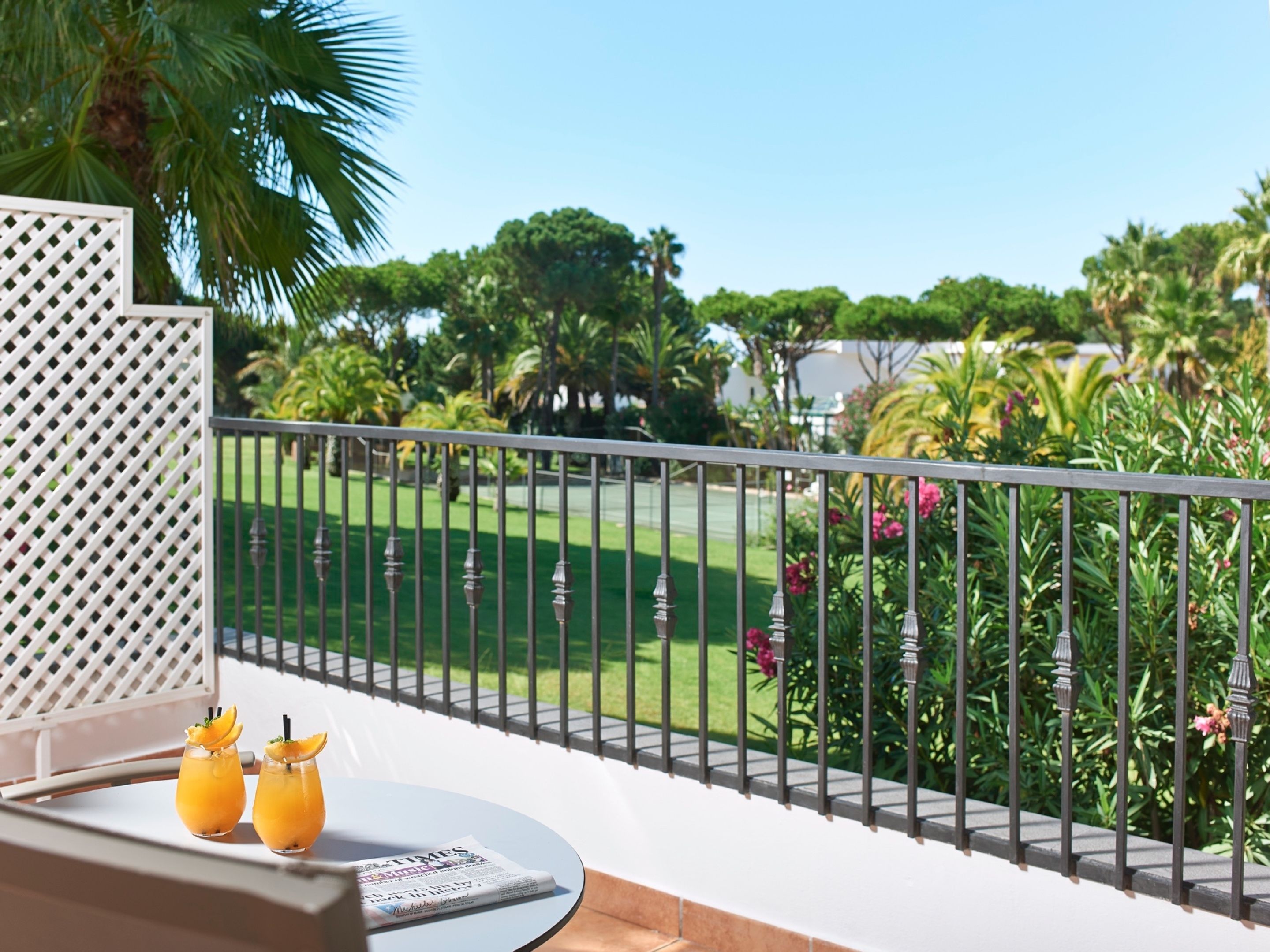 A balcony with a view of a well-maintained lawn and trees, two drinks on a tray, and a newspaper.