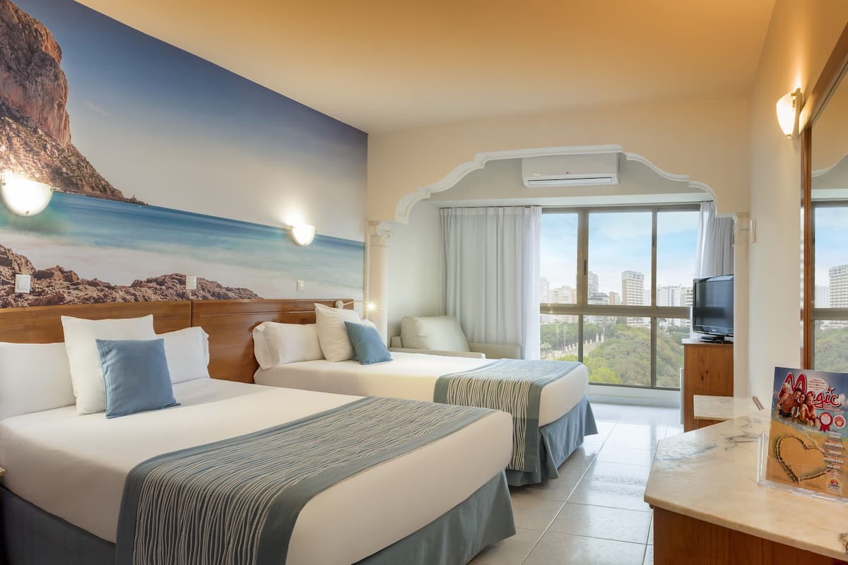 Habitación de hotel con dos camas, televisión, ventana grande con vista a la ciudad y un mural con una escena de playa.