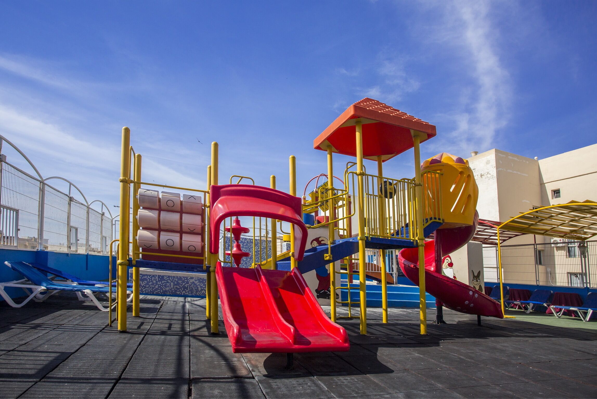 Un parque infantil con toboganes y estructuras para escalar, rodeado por una valla y un edificio al fondo.