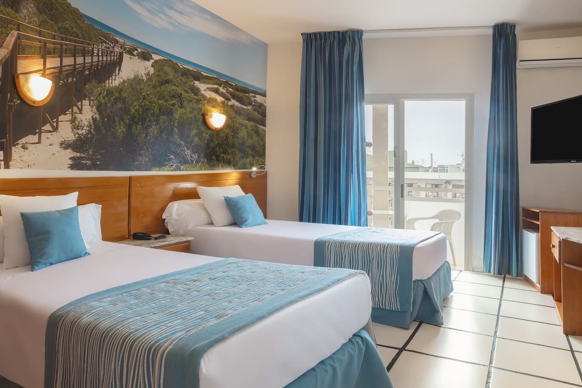 Habitación de hotel con dos camas, un ventanal grande, un televisor y un mural con una escena de playa.