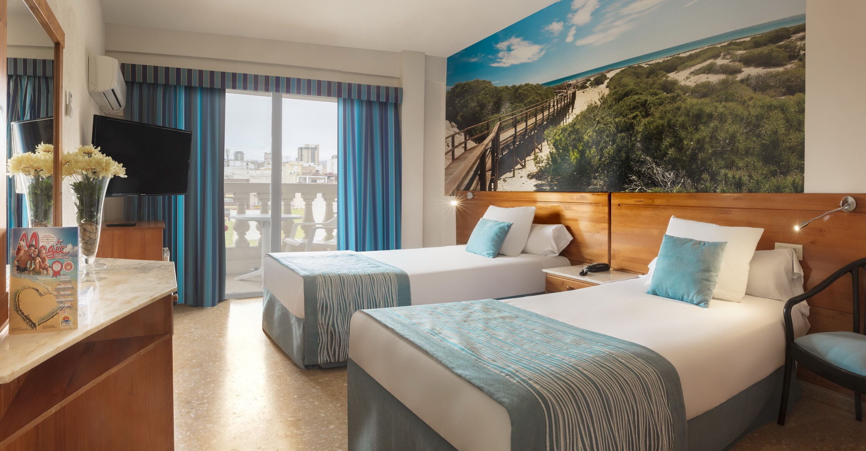 Habitación de hotel con dos camas, televisión, escritorio y un amplio mural de playa.