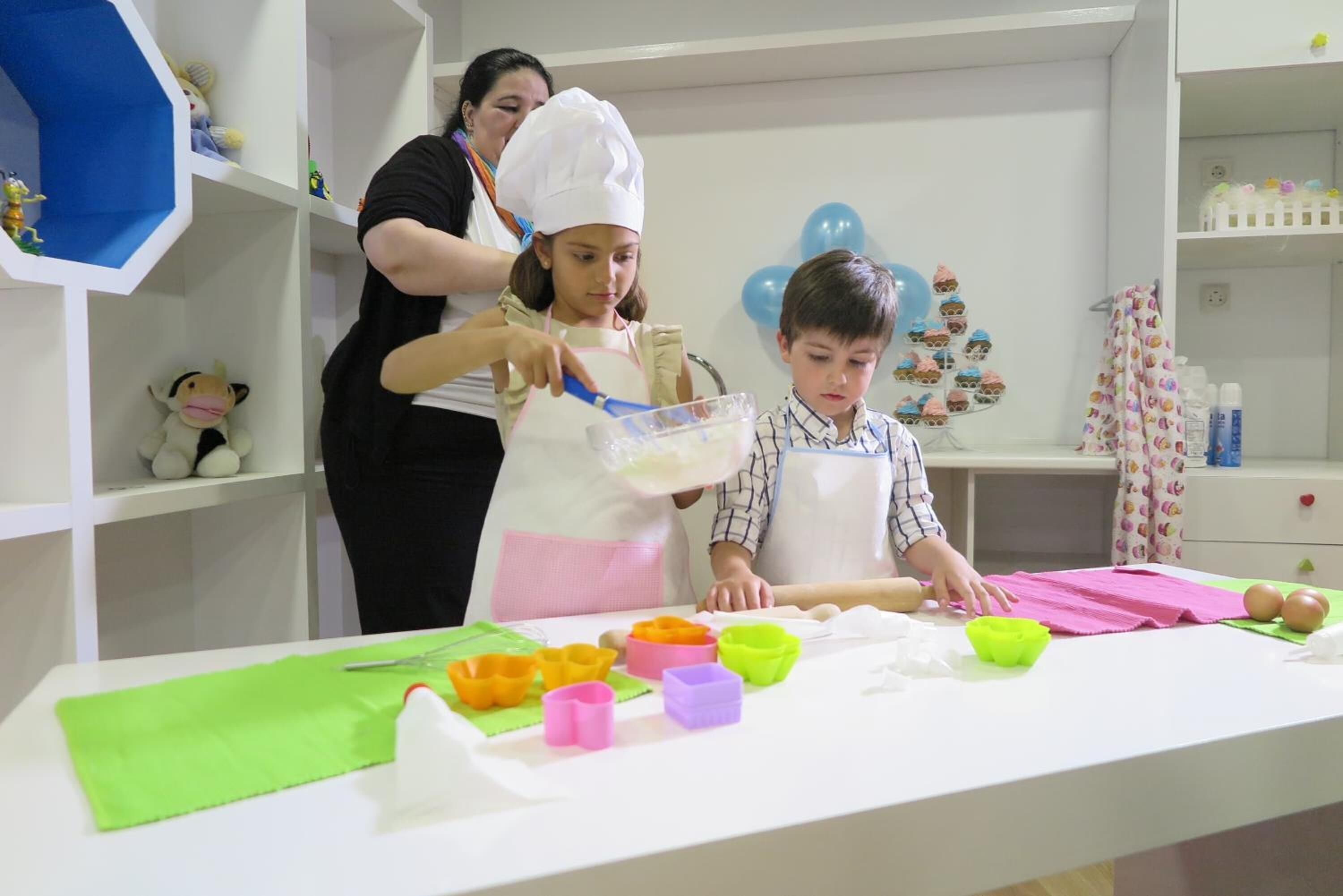 Una zona de cocina con una mesa preparada para una actividad de cocina, con varios moldes de repostería de colores y una persona con gorro de chef ayudando a un niño.