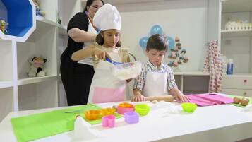 Una zona de cocina con una mesa preparada para una actividad de cocina, con varios moldes de repostería de colores y una persona con gorro de chef ayudando a un niño.