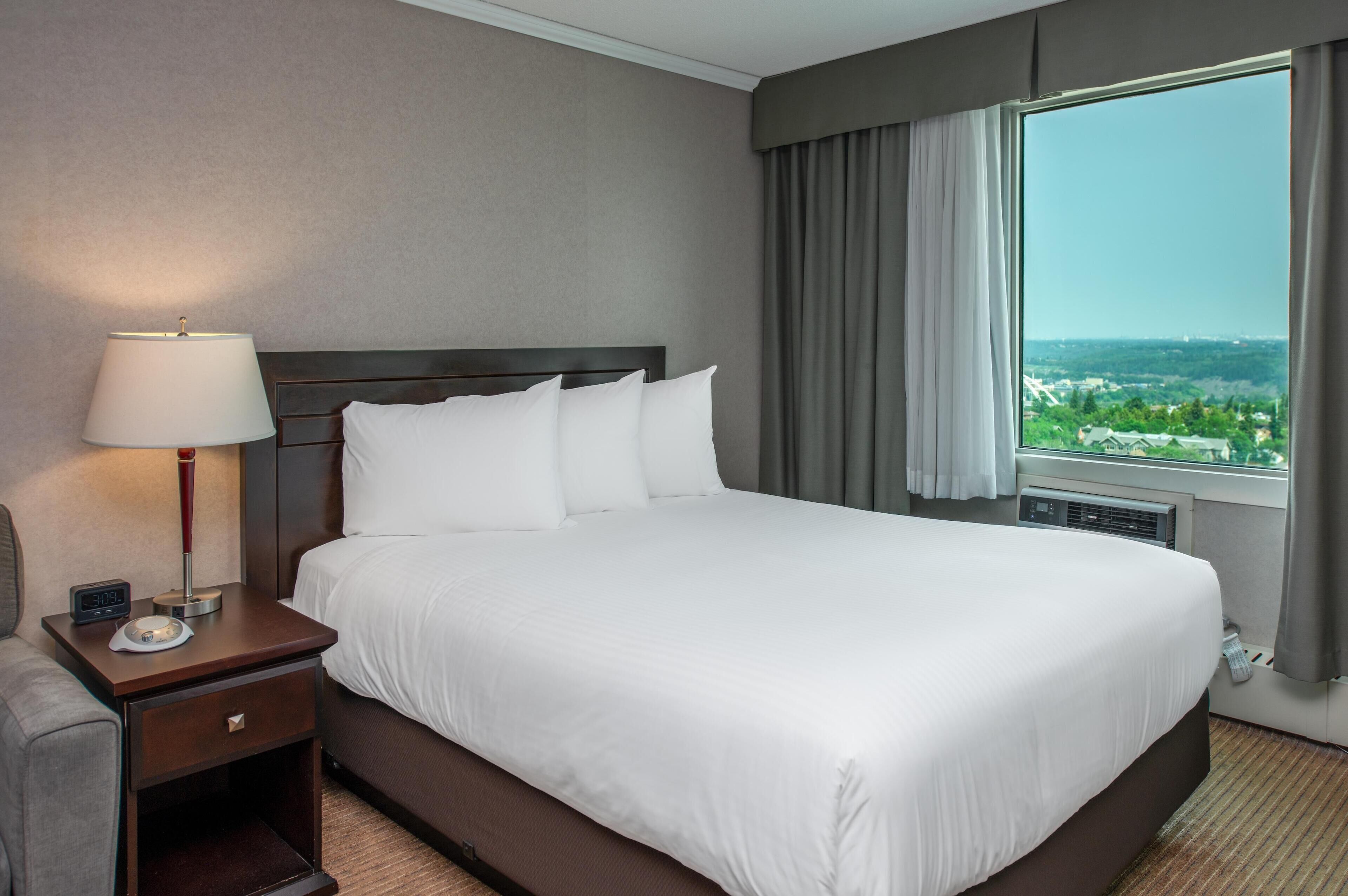 A hotel room with a large bed, a bedside table with a lamp, and a view of a cityscape through a window.