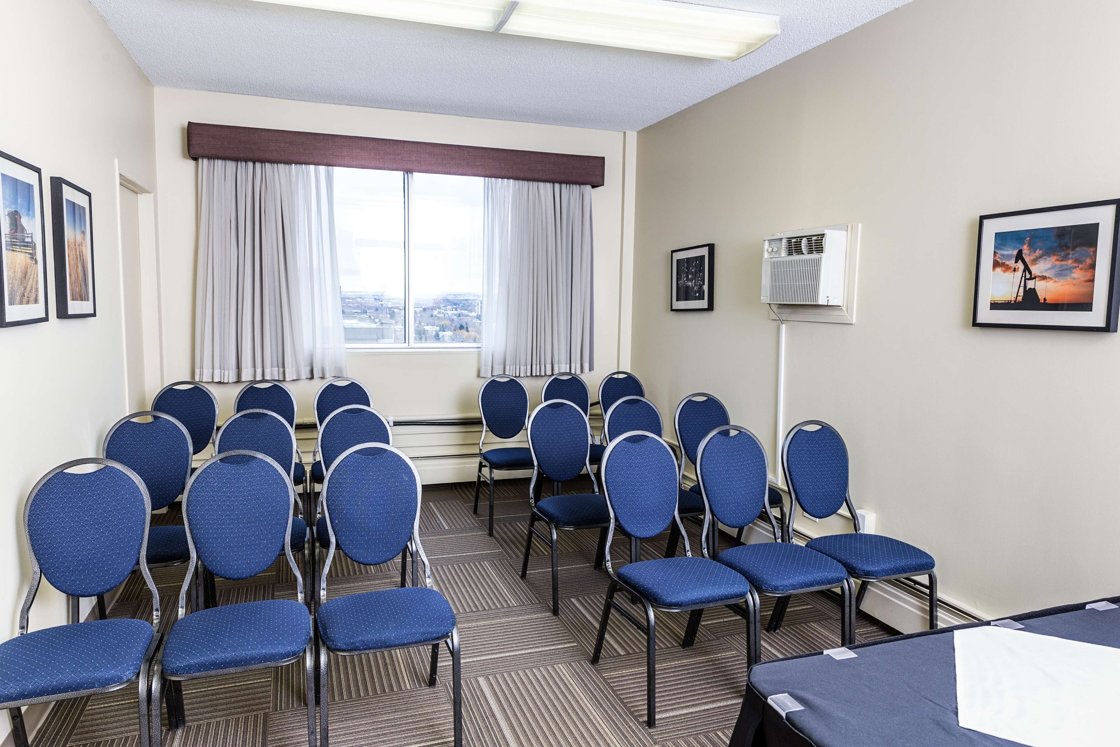 Une salle de conférence avec des rangées de chaises bleues, une grande fenêtre avec des rideaux et des tableaux encadrés accrochés aux murs.