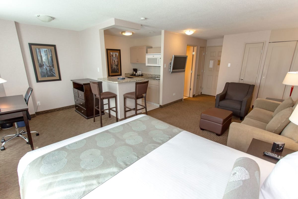 A hotel room with a large bed, a desk, a chair, a sofa, a kitchenette, and a bathroom.