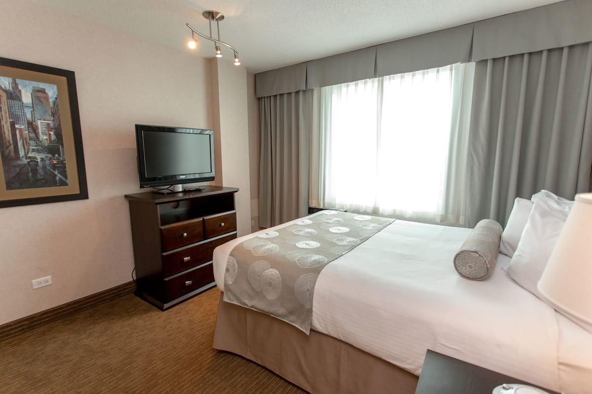 A hotel room with a bed, a television on a dresser, a window with curtains, and a painting on the wall.