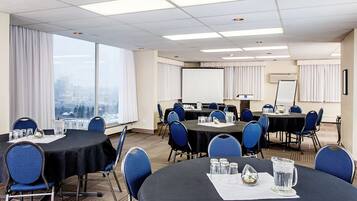 Uma sala de conferências com mesas redondas, cadeiras azuis, uma tela de projetor e vista para a paisagem urbana através de grandes janelas.