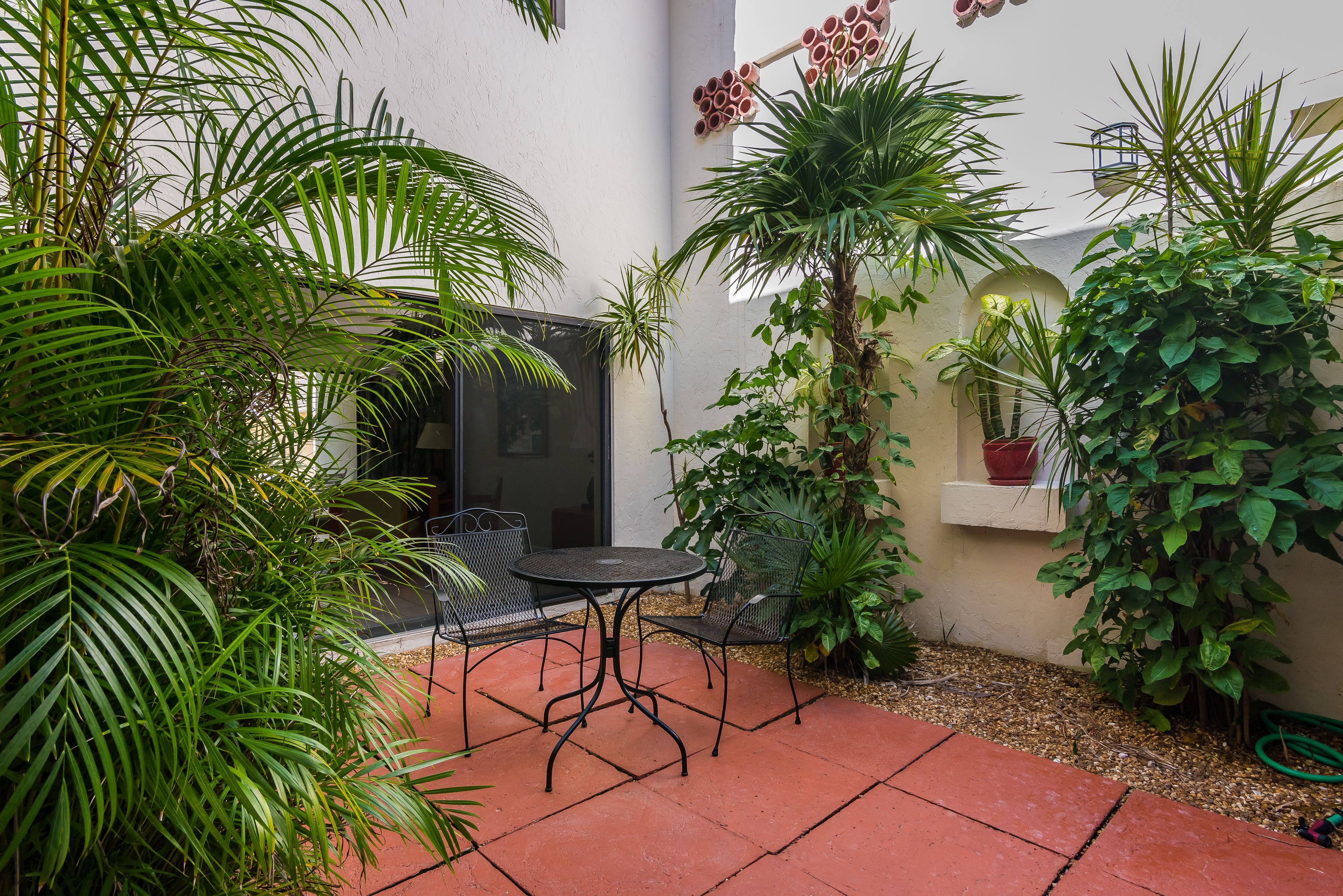 A patio with a table and chairs, surrounded by various plants and a white wall.