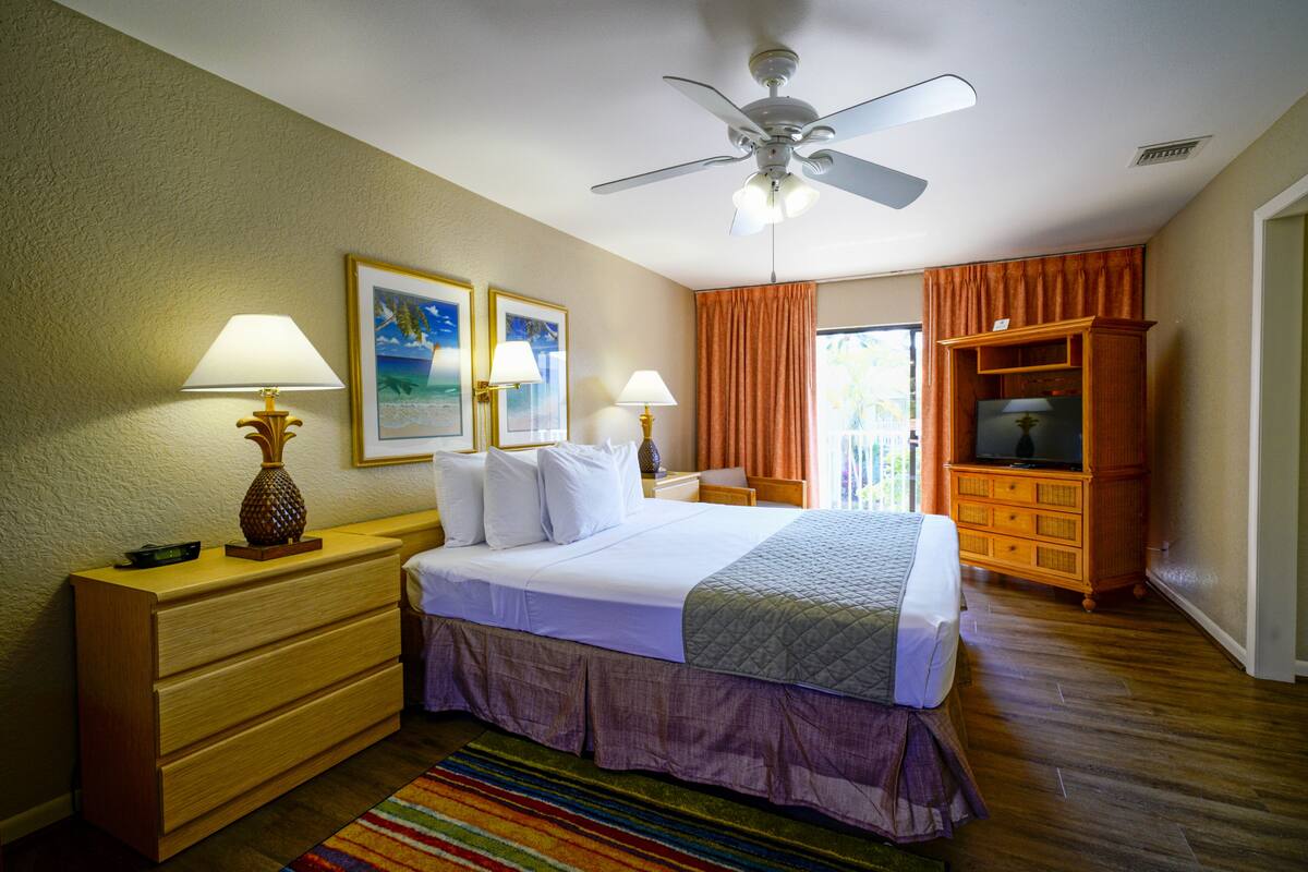 A hotel room with a bed, nightstands, a dresser, a ceiling fan, a TV, and a window with curtains.