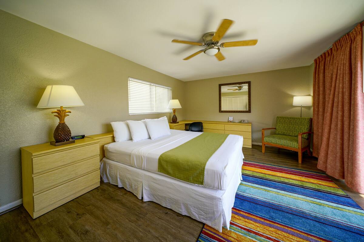 A hotel room with a bed, two bedside tables, a chair, a ceiling fan, a window with blinds, and a colorful striped rug.